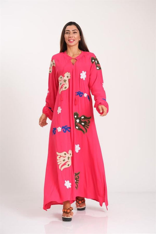A woman wears a long, pink, crew-neck viscose dress featuring colorful embroidery and tassels.