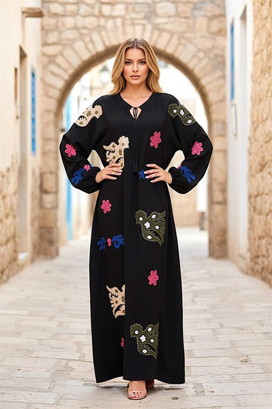 A woman wears a long-sleeved black maxi dress featuring colorful floral embroidery and tassels.