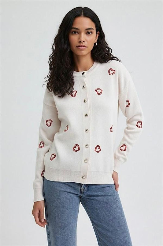 Kırmızı kalp nakışlı, düğmeli, beyaz triko hırka giyen bir kadın.