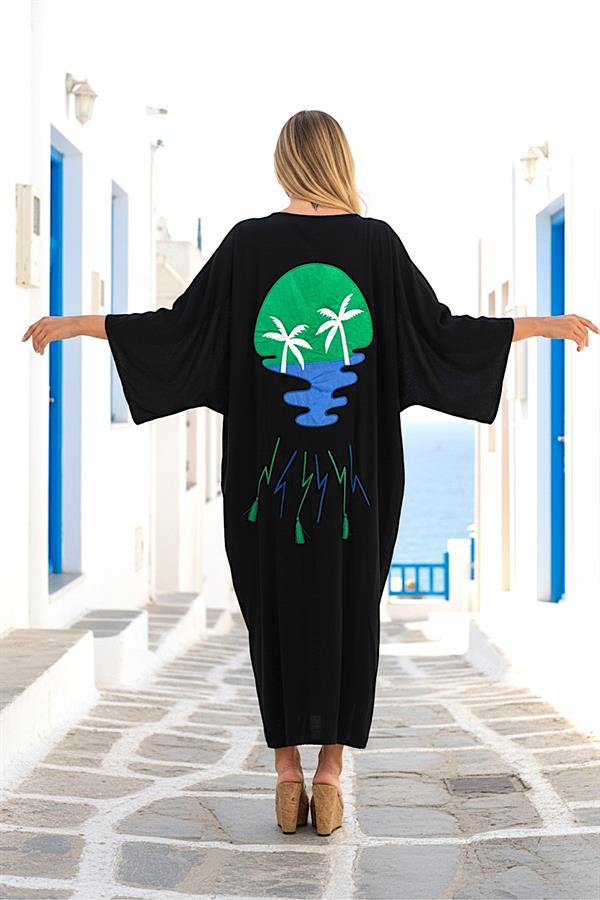 A woman wears a long black viscose shirt dress featuring a palm tree applique and tassels on the back.